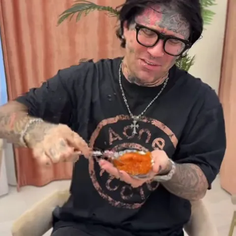 Мужчина с татуировками на лице и руках, одетый в черную футболку и очки, ест красную икру ложкой прямо из тарелки на руке.