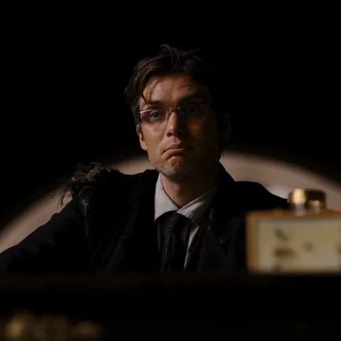 Мужчина в очках и костюме с серьезным выражением лица сидит за столом на темном фоне.