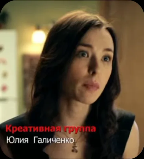 Брюнетка в чёрном топе с кулоном эмоционально разговаривает на фоне тёплого интерьера.
