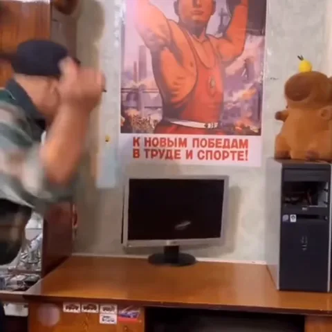 Человек в кепке разбивает топором монитор на столе, на стене плакат «К новым победам в труде и спорте!».