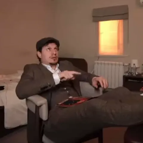 Сидящий мужчина в шляпе активно жестикулирует рукой, указывая пальцем.