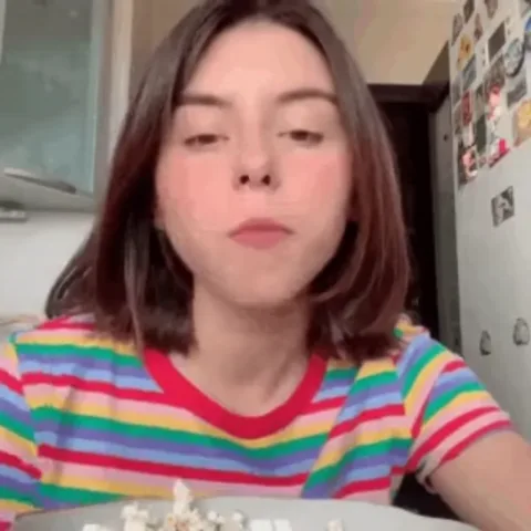 Брюнетка с коротким каре в радужной футболке сидит перед миской с попкорном, пережёвывает его и берёт следующий кусочек.