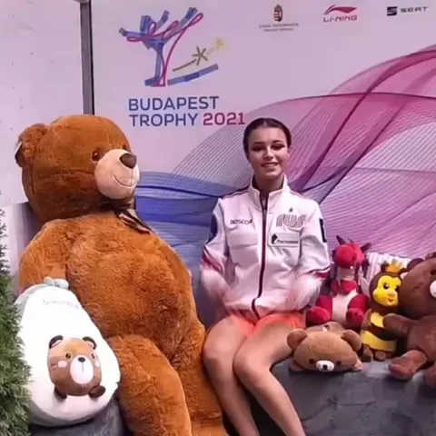 Девушка в спортивной куртке с надписью Россия делает жесты руками: машет и складывает пальцы в сердце. Вокруг неё большие плюшевые медведи.