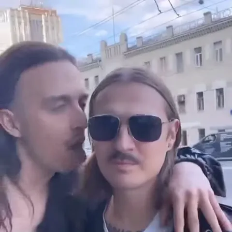 Двое мужчин на улице: один с длинными волосами целует другого в очках в щёку, обняв его за плечи.