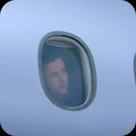 Молодой человек с тёмными волосами смотрит из окна самолёта и улыбается.