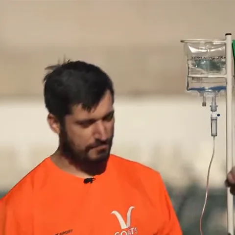 Мужчина в оранжевой футболке указывает пальцем на медицинскую капельницу.