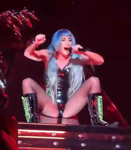 Lady Gaga в голубом парике и черном латексном боди поет в микрофон на концерте, подняв правую руку в жесте силы.
