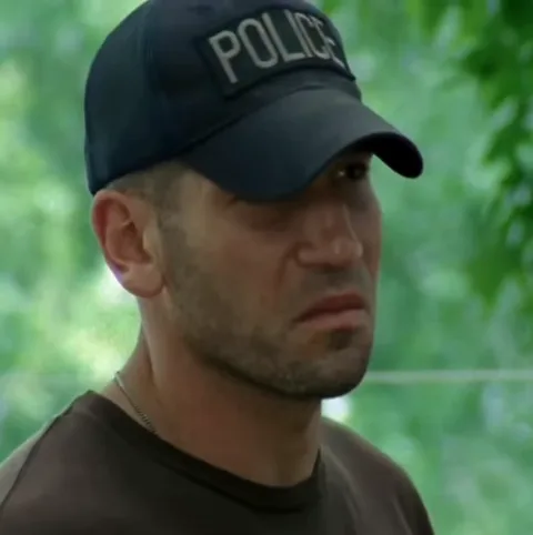 Мужчина в кепке с надписью POLICE внимательно смотрит в сторону на фоне зелени.