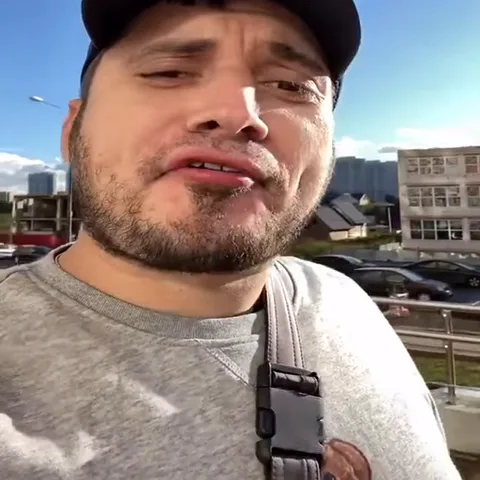 Мужчина в кепке и серой толстовке плюется белой пеной на камеру.