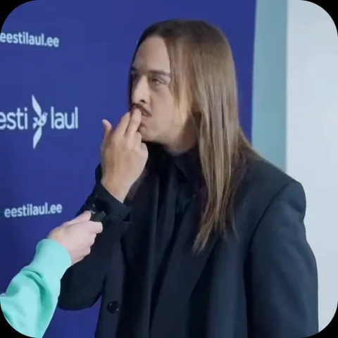 Мужчина с длинными светлыми волосами в черном пиджаке на фоне синей стены с логотипами прижимает палец к носу.