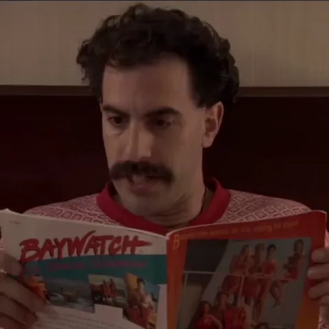 Борат держит в руках открытый журнал с надписью «Baywatch» и внимательно читает его.