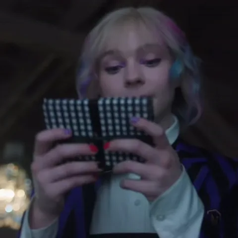 Анимация Энид Синклер из сериала «Уэнсдей», которая держит клетчатый блокнот и задумчиво смотрит вверх.