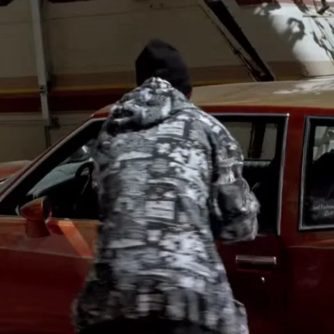 Человек в черно-белой куртке с узором и черной шапке забирается в красную машину через боковое окно.