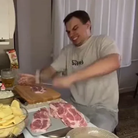 Мужчина в серой футболке энергично бьет кухонным молотком по мясу, лежащему на деревянной доске. На столе также видны сырая картошка и другие продукты.