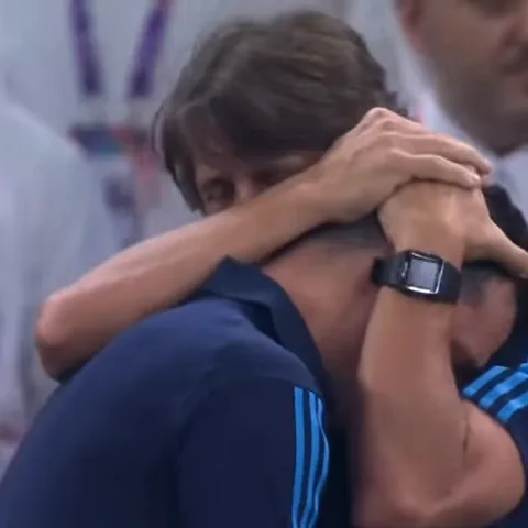 Мужчины в синих футболках с белыми полосами обнимают друг друга на стадионе.