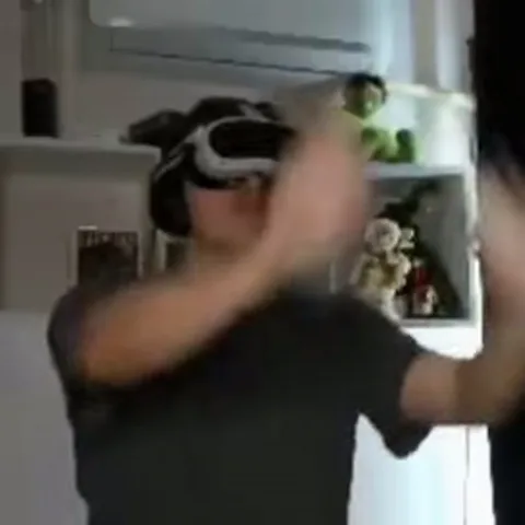 Человек в VR-шлеме и наушниках активно жестикулирует перед камерой на фоне полки с игрушками.