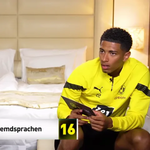 Молодой темнокожий мужчина в спортивной форме Баварии (BVB) сидит на кровати. Он держит карточку, читает её, удивляется, а потом закрывает глаза рукой в жесте «фейспалм».