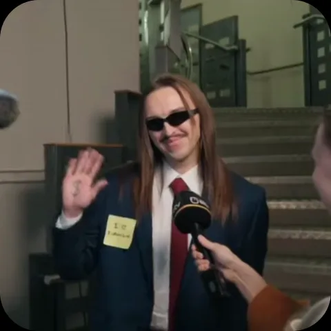 Мужчина с длинными волосами, в темных очках и деловом костюме, улыбается и поднимает руку в жесте приветствия перед микрофоном.
