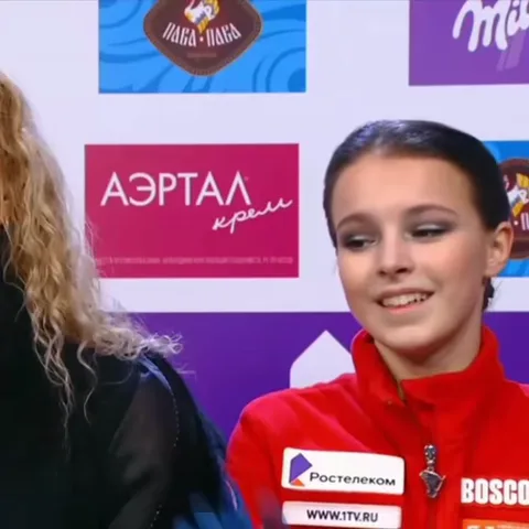 Спортивная девушка с темными волосами, собранными назад, делает жест «сердце» из рук в голубых перчатках на фоне пресс-волла.