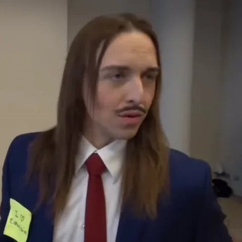 Длинноволосый мужчина в деловом костюме говорит перед камерой.