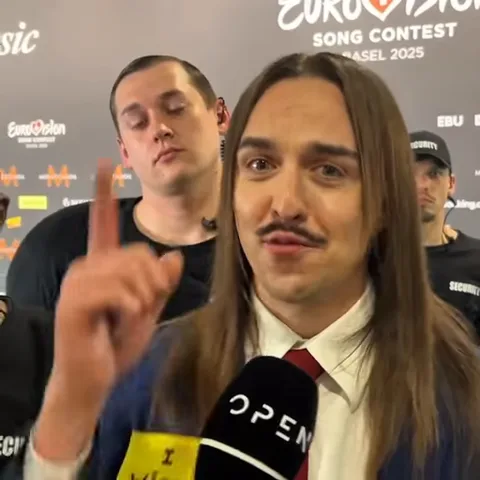 Tommy Cash с длинными волосами и усами в костюме говорит в микрофон во время пресс-конференции.