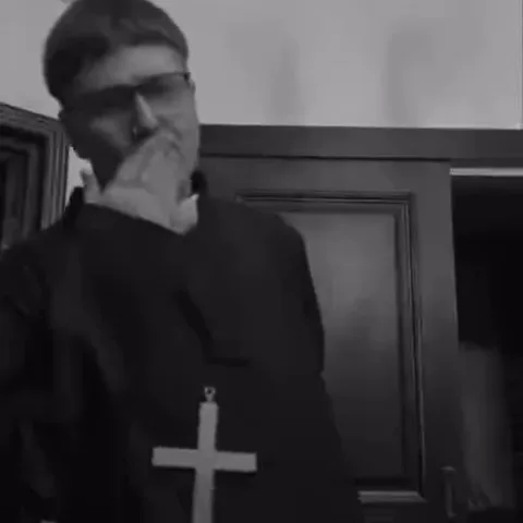 Мужчина в очках и одежде священника с большим крестом на груди реагирует на что-то: сначала закрывает рот от удивления, потом пожимает плечами.