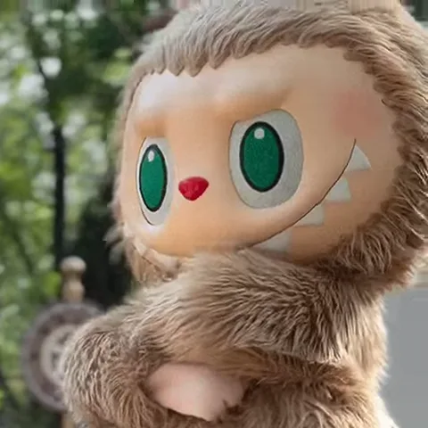 Крупный план плюшевой фигурки Labubu коричневого цвета со скрещенными на груди руками и зелеными глазами.