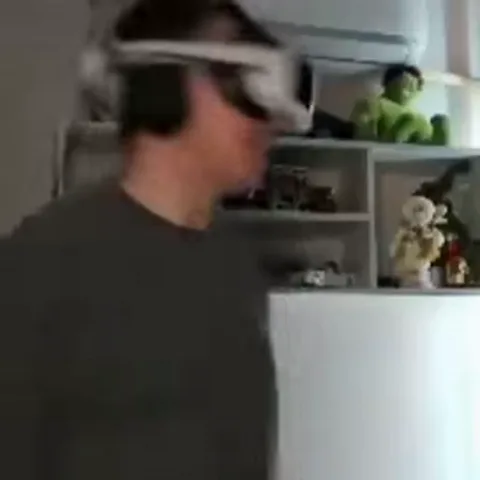 Человек в серой футболке и белых VR-очках быстро поворачивает голову из стороны в сторону на фоне полок с игрушками.
