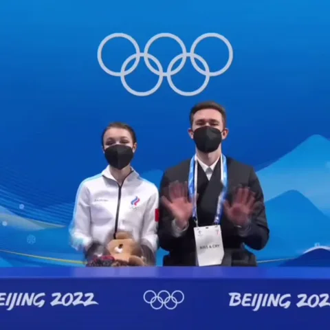 Люди в масках аплодируют на фоне логотипа зимних Олимпийских игр.
