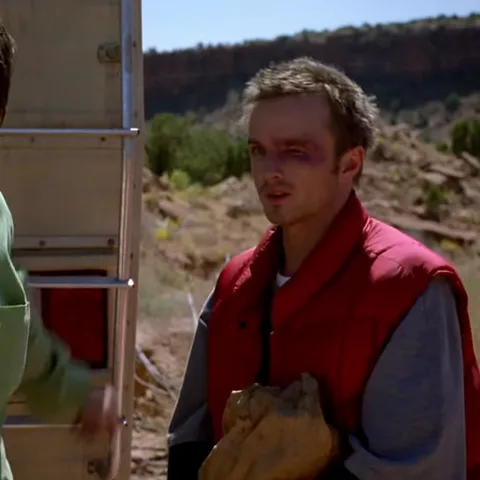 Молодой человек в красной жилетке с синяком под левым глазом на фоне пустыни, жестикулирует рукой.