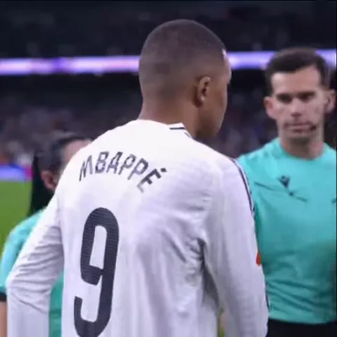 Вид со спины на футболиста в белой футболке с надписью MBAPPE и цифрой 9 на фоне стадиона.