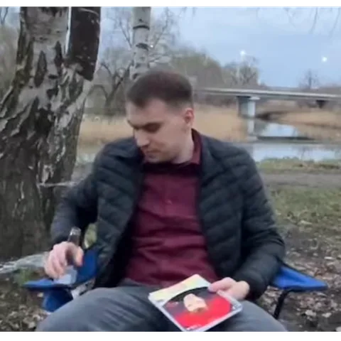 Мужчина сидит на улице у воды, держит в руках журнал и пьет из бутылки.