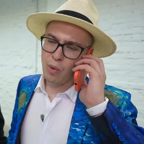Мужчина в шляпе и очках, в синем пиджаке с узором, держит оранжевый телефон у уха и активно жестикулирует, выражая разные эмоции.