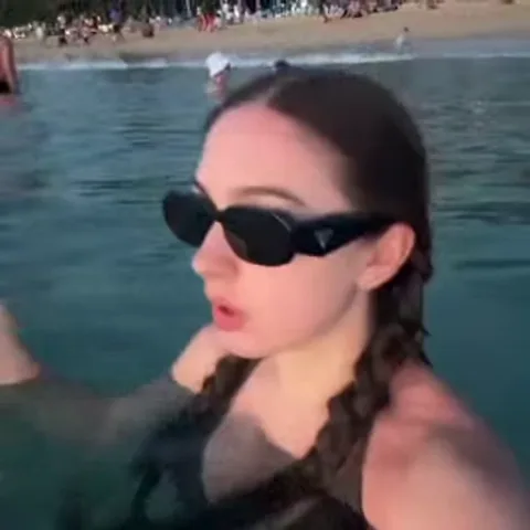 Блондинка с двумя длинными косами стоит по плечи в воде. На ней черные солнцезащитные очки, которые она рукой задвигает на макушку. На заднем плане виден берег с людьми.